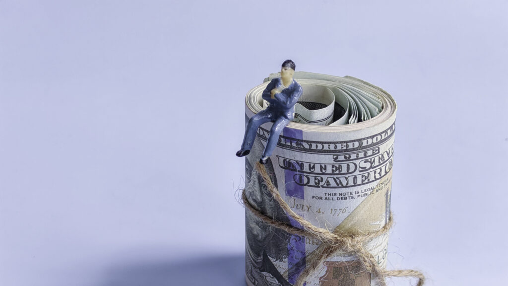 Miniature Businessman Sitting on Rolled US Dollar Bills Tied with Twine.